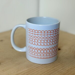 Könnte beinhalten: Eine hellblaue Keramiktasse mit weißem Henkel. Die Tasse ist mit einem sich wiederholenden Muster aus orangefarbenen geometrischen Formen verziert. Die Tasse steht auf einer Holzoberfläche.