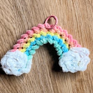 Könnte beinhalten: Ein regenbogenförmiges Bastelobjekt aus flauschigen Pfeifenreinigern in Rosa, Gelb und Blau, mit weißen Pfeifenreiniger-Wolken an beiden Enden. Ein kleiner rosa Kunststoffring ist oben in der Mitte der gewölbten Regenbogenstruktur befestigt.