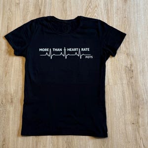 Könnte beinhalten: Schwarzes T-Shirt mit weißem Text und einer Herzschlag-Grafik. Der Text lautet "MORE THAN HEART RATE POTS". Das Shirt besteht aus einem weichen Material und eignet sich für Freizeitkleidung.