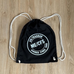 Op de afbeelding: Zwarte trekkoordtas met witte touwriemen. De tas heeft een witte cirkelvormige afbeelding met de tekst "UN-TRAGBAR ME/CFS IGNORED SINCE 1969". De tas is gemaakt van een duurzame stof en is geschikt voor het dragen van diverse items.