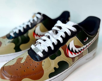 May include: A pair of custom Air Force 1 sneakers with a camouflage pattern in shades of brown, green, and tan. The shoes feature a shark-inspired design with white, red, and black accents, and white laces.