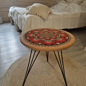 May include: A round wooden side table with a mandala design on the tabletop. The mandala features red, orange, and black colors. The table has black hairpin legs and sits on a beige rug. The table is approximately 18 inches in diameter.