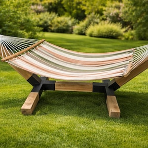 May include: A striped hammock with a wooden frame and black supports is set up on a grassy lawn. The hammock's fabric features horizontal stripes in shades of beige, green, and orange. The wooden frame is angled for stability.