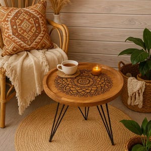 Könnte beinhalten: Ein runder Beistelltisch aus Holz mit Mandala-Design, getragen von schwarzen Metallbeinen, steht auf einem Jute-Teppich. Eine Tasse Tee, eine Kerze und ein gewebtes Kissen auf einem Rattanstuhl vervollständigen die gemütliche, Boho-Szene.