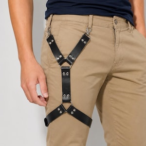 May include: A black leather leg harness featuring two top clips that attach to belt loops, connected by silver-toned metal rivets and rings. The harness forms a Y-shape that extends down to a horizontal strap encircling the upper thigh area.