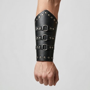 May include: Black leather arm guard with three adjustable straps and silver buckles. The arm guard is decorated with silver rivets along the edges. The product is designed to protect the forearm.