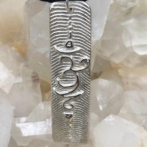 Puede incluir: Colgante de plata con un símbolo Om grabado en él. El colgante es rectangular y tiene una superficie texturizada.