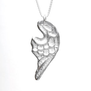 Puede incluir: Un colgante plateado con forma de ala estilizada, suspendido de una delicada cadena. El diseño del ala presenta texturas y patrones detallados, creando un efecto tridimensional. La joya se presenta sobre un fondo blanco liso.
