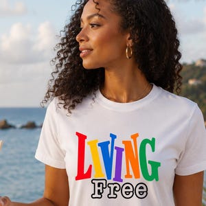 Puede incluir: Una camiseta blanca de manga corta con el texto "LIVING" en letras de bloque multicolores sobre la palabra "Free" en una fuente negra con contorno. La prenda tiene cuello redondo y un corte estándar con un estampado gráfico visible.
