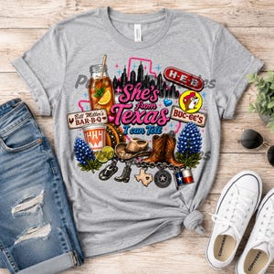 Può includere: Una t-shirt grigio melange con una stampa grafica che riporta il testo She's from Texas I can Tell. Il design include uno skyline, un bicchiere di tè freddo, un cappello da cowboy, stivali, lupini e vari loghi e icone a tema Texas.