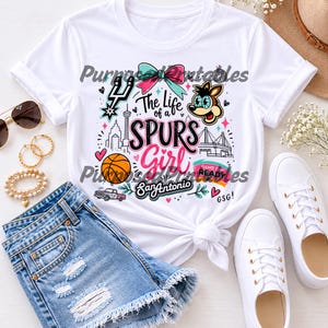 May include: A white short-sleeved t-shirt featuring a colorful graphic print with the text The Life of a SPURS Girl San Antonio GSG. The design includes a basketball, a bow, a cartoon character, a city skyline, and a pickup truck in pink and teal.