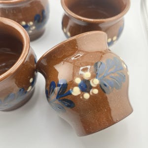 Może przedstawiać: Kolekcja małych, brązowych ceramicznych doniczek z błyszczącym wykończeniem. Każda doniczka ozdobiona jest ręcznie malowanym kwiatowym wzorem w kolorze niebieskim, białym i żółtym. Doniczki mają lekko rozszerzoną krawędź i rustykalny wygląd.