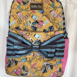 May include: A quilted backpack with a mustard yellow background featuring a pattern of eyeglasses, pencils, notebooks, and sneakers. The backpack has a large blue and gray striped bow, pink side panels, and the text "Matilda Jane".