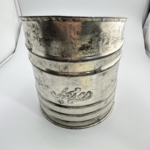 May include: A weathered, cylindrical metal container with a distressed silver finish. The container has horizontal bands and the embossed word "Asco" on the front. The vintage bucket has a slightly flared top.