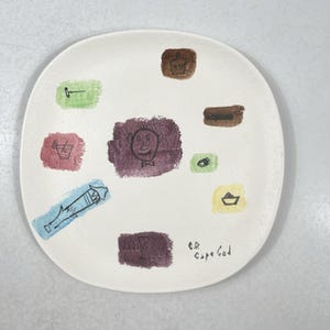 Könnte beinhalten: Cremefarbene Keramikplatte mit skurrilen Zeichnungen in verschiedenen Farben. Die Platte hat ein zentrales lila Quadrat mit einem lächelnden Gesicht, umgeben von anderen bunten Formen und Zeichnungen. Der Text "E.D. Cape Cod" ist auf der Platte geschrieben.