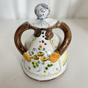 May include: A ceramic figurine of a woman in a white dress with brown arms and a head covering. The dress is decorated with yellow flowers and green leaves. The figurine has a bell-shaped body and is painted in a folk art style.