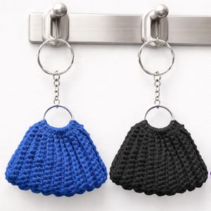 May include: Three handmade crochet keychains in blue, black, and purple, each with a circular metal ring. The keychains are hanging from a silver metal hook rack against a white background. The crochet design resembles a small skirt.