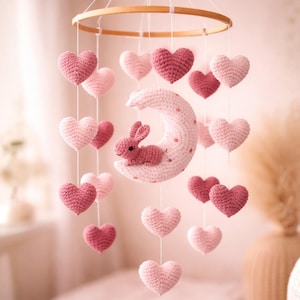 May include: A handmade baby mobile featuring a wooden ring with hanging crochet hearts in shades of pink and a crochet moon with a bunny. The mobile is suspended from a white string, creating a whimsical nursery decoration.