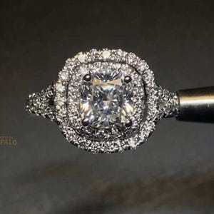 Puede incluir: Un anillo de tono plateado con una piedra preciosa central transparente de corte cojín rodeada por un doble halo de piedras transparentes más pequeñas de corte redondo. La banda está dividida y engastada con piedras adicionales. El texto OPALO es visible.