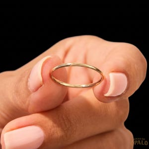 May include: A thin, polished gold band ring with a smooth, circular profile. The metal has a reflective, warm yellow tone and a simple, unadorned design. The ring is held in a position that highlights its uniform thickness and rounded shape.
