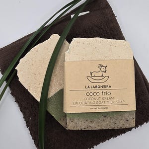 May include: A bar of exfoliating goat milk soap with a coconut cream scent, labeled "Coco Frio" by La Jabonera. The soap is layered with green and beige tones, resting on a brown washcloth, with green leaves. Net wt 5 oz (141g).