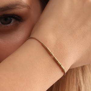 May include: A delicate gold bracelet with a linear design. The bracelet features a thin chain with several cylindrical and textured gold beads. The bracelet is worn on the wrist.