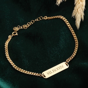 May include: Gold-colored chain bracelet with a rectangular plate engraved with Roman numerals. The bracelet is displayed on a dark green velvet surface, with a sprig of dried flowers in the background. The bracelet's chain is made of small, interconnected links.