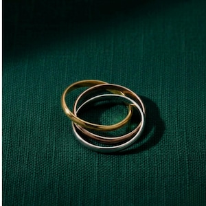 Può includere: Tre anelli intrecciati in oro, oro rosa e argento poggiano su una superficie strutturata verde scuro. Gli anelli sono lisci e lucenti, creando un design semplice ma elegante. Gli anelli sono un classico gioiello.