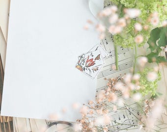 Pode incluir: Uma folha de papel branca em branco com um selo floral, tesouras e partituras, dispostas numa bandeja de vime. A cena é decorada com fita branca, flores secas e folhagem verde, criando uma estética vintage.