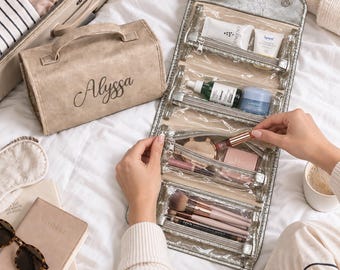 May include: A beige travel toiletry bag with the name Alyssa printed in black script, featuring a matching metallic silver roll-up organizer with four clear zippered compartments filled with various skincare, makeup products, and cosmetic brushes.