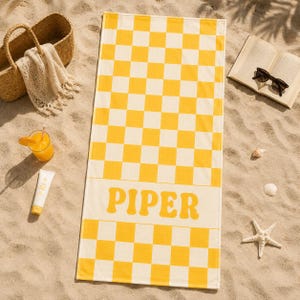 May include: A rectangular beach towel featuring a yellow and white checkered pattern with a solid horizontal band across the center containing the name PIPER in bold yellow letters. The towel is laid flat on a sandy surface.