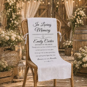 May include: A white fabric chair sash with the text "In Loving Memory" and "Emily Carter, Mother of the Bride" in elegant script. The sash is tied to a wicker chair, with a wedding theme.