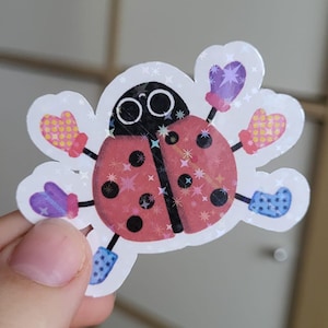 May include: A whimsical sticker featuring a cartoon ladybug with a red body, black spots, and large black eyes. The ladybug has arms and legs with colorful mittens and boots. The sticker has a white border and a sparkly finish.