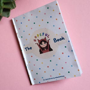 Può includere: Un piccolo libro intitolato "The Hopeful Book" con un'illustrazione a cartoni animati di una ragazza con un mantello rosso. La copertina è bianca con pois colorati. Una pianta succulenta e un vaso a forma di orso sono sullo sfondo.