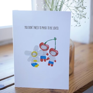Può includere: Biglietto di auguri bianco con la scritta "YOU DON'T NEED TO MASK TO BE LOVED." con illustrazioni di un fiore e due ciliegie, tutte con facce sorridenti e pantaloncini blu. Il biglietto è su una superficie di legno.