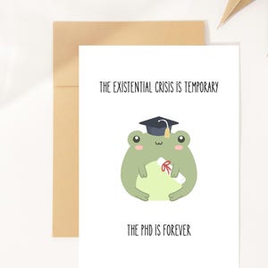 Könnte beinhalten: Eine weiße Grußkarte mit einem Cartoon-Frosch, der einen Abschluss-Hut trägt und eine Urkunde hält. Der Text auf der Karte lautet: "THE EXISTENTIAL CRISIS IS TEMPORARY" und "THE PHD IS FOREVER". Die Karte liegt in einem braunen Umschlag.