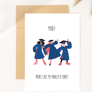 Puede incluir: Una tarjeta de felicitación blanca con el texto "PHD? MORE LIKE PH-INALLY D-ONE!". Presenta ilustraciones de tres graduados con túnicas azules y birretes. La tarjeta va acompañada de un sobre marrón y una hoja de palma seca.
