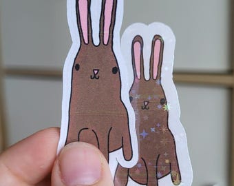 May include: Two brown bunny stickers with pink inner ears and a white outline. The larger bunny has a simple design, while the smaller one has a sparkly, glittery effect. The stickers are held in a hand.