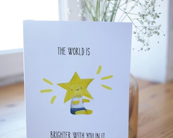 May include: A white greeting card with the text "THE WORLD IS BRIGHTER WITH YOU IN IT." features a yellow star character with a smiling face. The star is wearing a blue and white outfit and has yellow rays emanating from it.