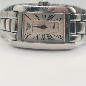 May include: Silver-toned rectangular wristwatch with a light pink face. The watch features Roman numeral hour markers, a subdial, and the brand name "Emporio Armani". The watch band is made of interlocking silver links.