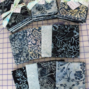 May include: Stacks of fabric squares in various shades of blue, featuring floral and leaf patterns. Some stacks are tied with ribbons and tags. The fabrics have a batik-like appearance, with dark and light blue hues creating contrast.