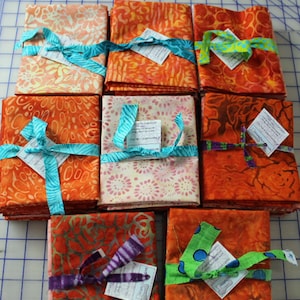 May include: Nine fabric bundles in shades of orange, pink, and cream, each tied with a colorful ribbon. The fabrics feature various patterns, including floral and abstract designs. Small tags are attached to each bundle.