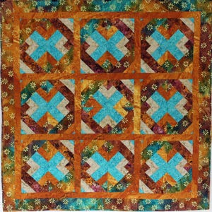 May include: A handmade quilt featuring a repeating pattern of turquoise crosses within square blocks. The blocks are framed by orange and brown borders, with a floral patterned edge. The quilt is a mix of warm and cool colors.