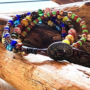 May include: A multi-strand beaded bracelet with a leather strap and a decorative metal clasp. The bracelet features colorful beads in shades of blue, green, yellow, red, and brown. The bracelet is displayed on a piece of driftwood.