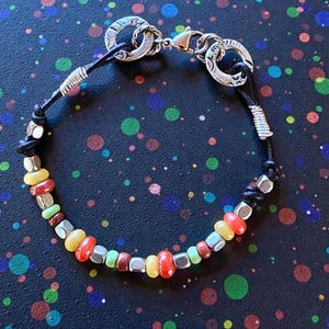 May include: A handcrafted bracelet featuring a black leather cord and a variety of colorful beads. The beads include red, yellow, green, and silver tones. The bracelet has a silver clasp and decorative silver rings.