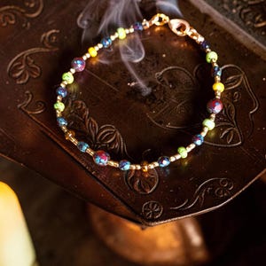 May include: A beaded bracelet with a gold clasp and small gold beads. The bracelet features colorful beads in shades of blue, green, yellow, and red. The bracelet is resting on a dark, ornate surface.