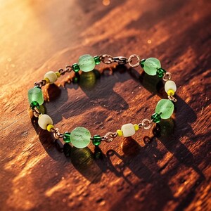 May include: A delicate bracelet featuring a series of light green, round beads interspersed with smaller green, white, and yellow beads. The bracelet is connected by a gold-colored chain and clasp, creating a simple yet elegant design.