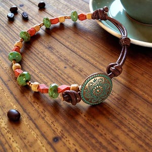 May include: A handcrafted bracelet featuring a mix of beads in green, orange, and beige, connected by a brown leather cord. The bracelet includes a decorative, oval-shaped, turquoise-colored metal piece with an embossed floral design.