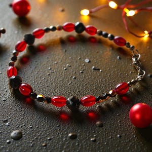 May include: A beaded bracelet featuring red and black beads, with a silver clasp. The bracelet is set against a dark, textured surface, with small red spheres and warm-toned fairy lights in the background.