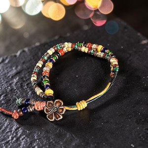 May include: A multi-strand bracelet with colorful beads in shades of red, yellow, green, and blue. The bracelet features a bronze flower charm and a knotted closure. The bracelet is on a dark surface.
